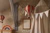 A cozy children's room features floral wallpaper, a burgundy hot air balloon decor, a framed alphabet chart, a fabric canopy, white pennant banner, and a colorful rainbow nightlight on a rattan cabinet.