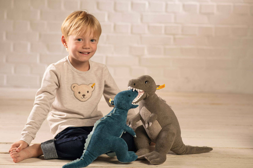 A young blond boy in a beige shirt sits on a light wooden floor, smiling and holding two plush toys—a Steiff Thaisen Tyrannosaurus "T-Rex" Dinosaur Plush Toy, 14 Inches, and another dinosaur—facing each other as if playing.