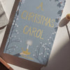 The "A Christmas Carol | Collector's Edition| Hardcover Book" rests on a white surface in sunlight, its blue cover adorned with gold lettering, holly accents, and a candle—evoking Ebenezer Scrooge’s classic tale.