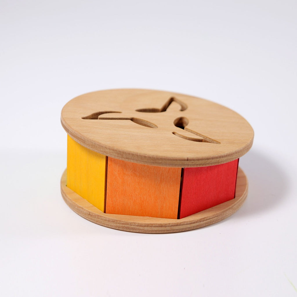 Grimm's Sound and Color Wheel is a non-toxic wooden toy with a round base and top, featuring three colorful sides—yellow, orange, and red—and three cutouts on top to help develop your baby’s motor skills.