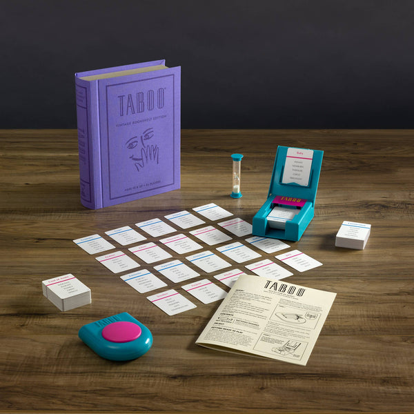 The Board Game: Taboo Vintage Bookshelf Edition is displayed on a wooden table with a purple box, cards, hourglass timer, blue buzzer with pink button, card holder, instruction sheet, and organized card stacks.