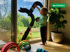 A young child plays with the Large Flexible Toy Road Set - Highway (24-Piece) on a glass door. Colorful wooden arches are on the floor, a large potted plant is nearby, and a "Flexible" label appears in the upper right corner.