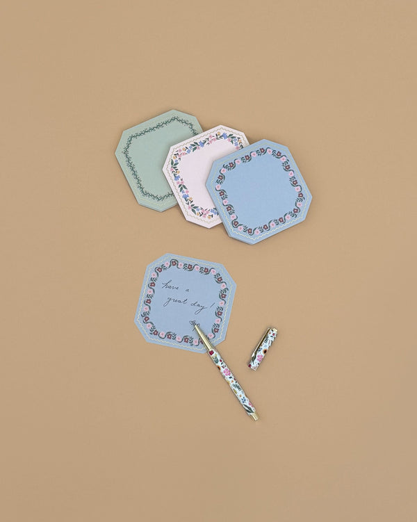 Three decorative, octagonal Gemma Mini Notepad Trio notepads with gold details and floral borders are displayed on a beige surface—one features the handwritten message “have a great day,” with a floral-patterned pen beside it.