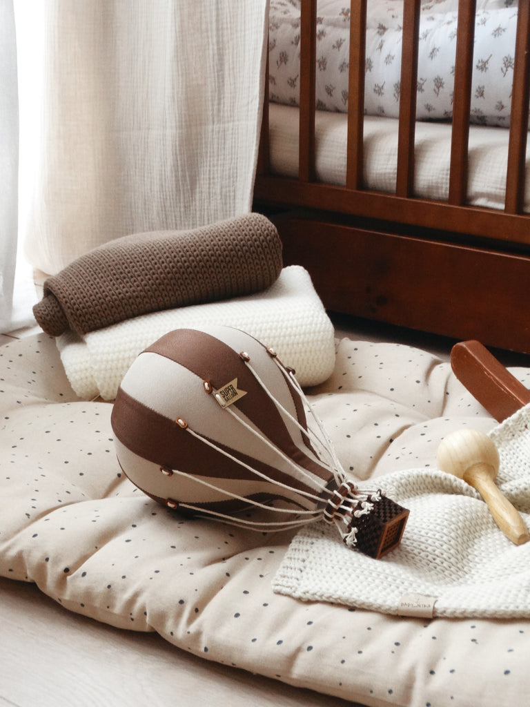 A soft, neutral nursery featuring a crib, folded blankets, polka-dot mat, decorative Hot Air Balloon - Light Brown / Dark Brown toy, and a wooden rattle arranged on the floor for a charming children’s room look.