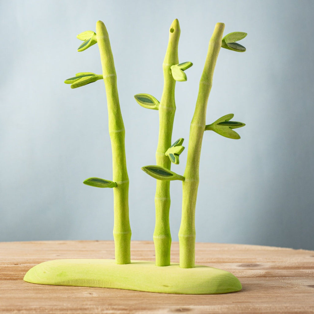 The Bumbu Handmade Wooden Red Panda Family with Bamboo Set features three stylized green bamboo stalks with leaves on a matching base—an ideal Montessori learning toy accent displayed on a wooden surface against a light blue background.