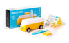 A VW Beach Bus Sunset toy van in yellow and white with a blue surfboard, yellow roof box, magnetic camper top, sticker sheet, and "BEACH BUS" retail box is displayed on a white background.