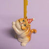 The Exotic Shorthair Cat Cake Topper features a small ceramic cat with orange stripes and a red-and-white party hat holding a yellow candle, perfect for cat-themed parties. Displayed on a light purple background.