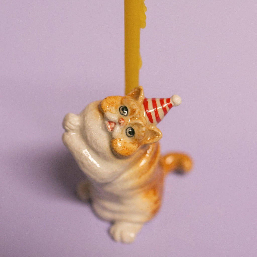 The Exotic Shorthair Cat Cake Topper features a small ceramic cat with orange stripes and a red-and-white party hat holding a yellow candle, perfect for cat-themed parties. Displayed on a light purple background.