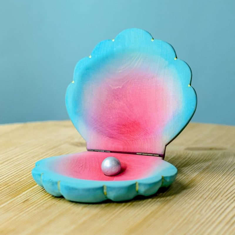 A small, shiny pearl rests inside the Bumbu Handmade Wooden Big Shell with Pearl—an open blue and pink clam shell, handcrafted and displayed on a light wooden surface against a soft blue background.