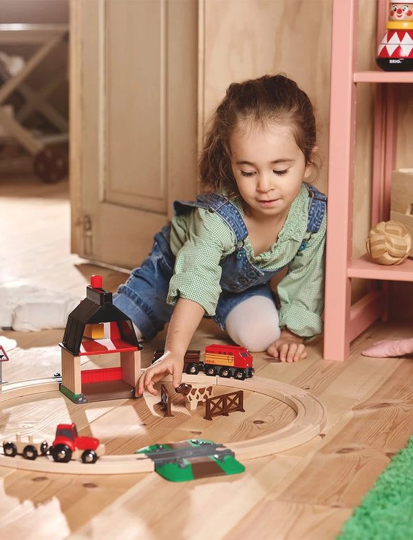 A young girl in a green shirt and denim overalls plays on a wooden floor with the BRIO World Train Set Farm Railway Set, arranging its magnetic train, barn, tractor, and animal figures as she focuses in her cozy room.