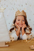 A smiling young girl wears the Crown - Gold Sequins, resting her chin on her hands, surrounded by warm fairy lights and gold star decorations, with a white cone tree in the foreground.