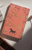 The "Black Beauty | Collector's Edition | Hardcover" features a pink and gold cover with a black horse silhouette and floral accents, resting on a white surface as sunlight casts gentle shadows across its elegant design.