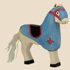 The Holztiger Tournament Horse, Blue, is a handcrafted wooden toy horse featuring a blue body, red and black harness, black hooves, and a white mane and tail. Ships in one week. Set against a plain beige background.