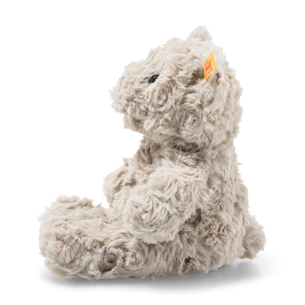 The Steiff Honey Teddy Bear Stuffed Plush, 8 inches, is a soft, light brown bear with curly fur, dark eyes, and an ear tag. It sits upright in profile facing left against a white background—a classic children’s toy.
