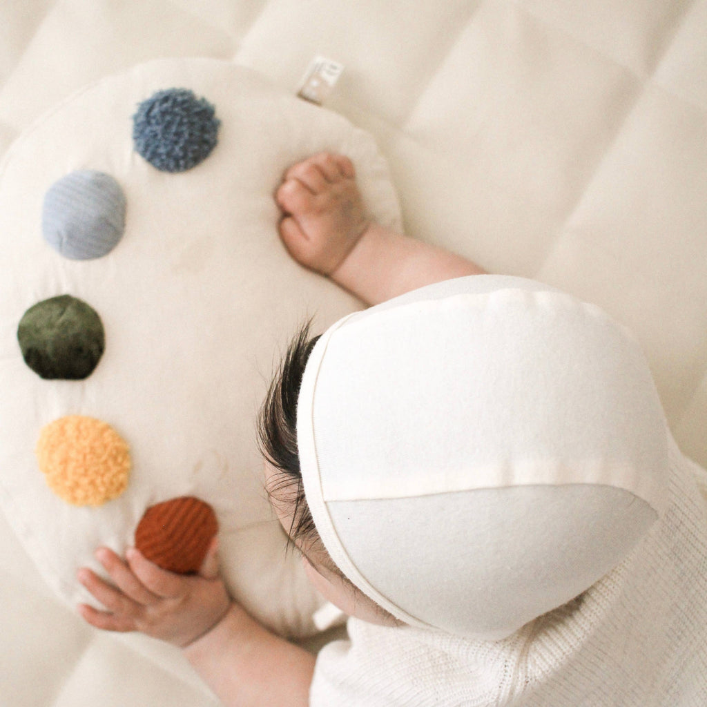 A baby wearing a cream cap touches the Tummy Time Palette, a soft, cream tummy time pillow adorned with six colorful pom-poms in a curved design on a light surface.