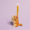 The Exotic Shorthair Cat Cake Topper is a small ceramic figurine of a cat in a red and white party hat holding a lit yellow candle, ideal for cake decoration or cat-themed parties, displayed on a light purple background.