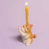 The Exotic Shorthair Cat Cake Topper features a ceramic cat figurine in a red-striped party hat, balancing a lit yellow candle on its back—ideal for cat-themed parties and birthdays. Set against a light purple background.