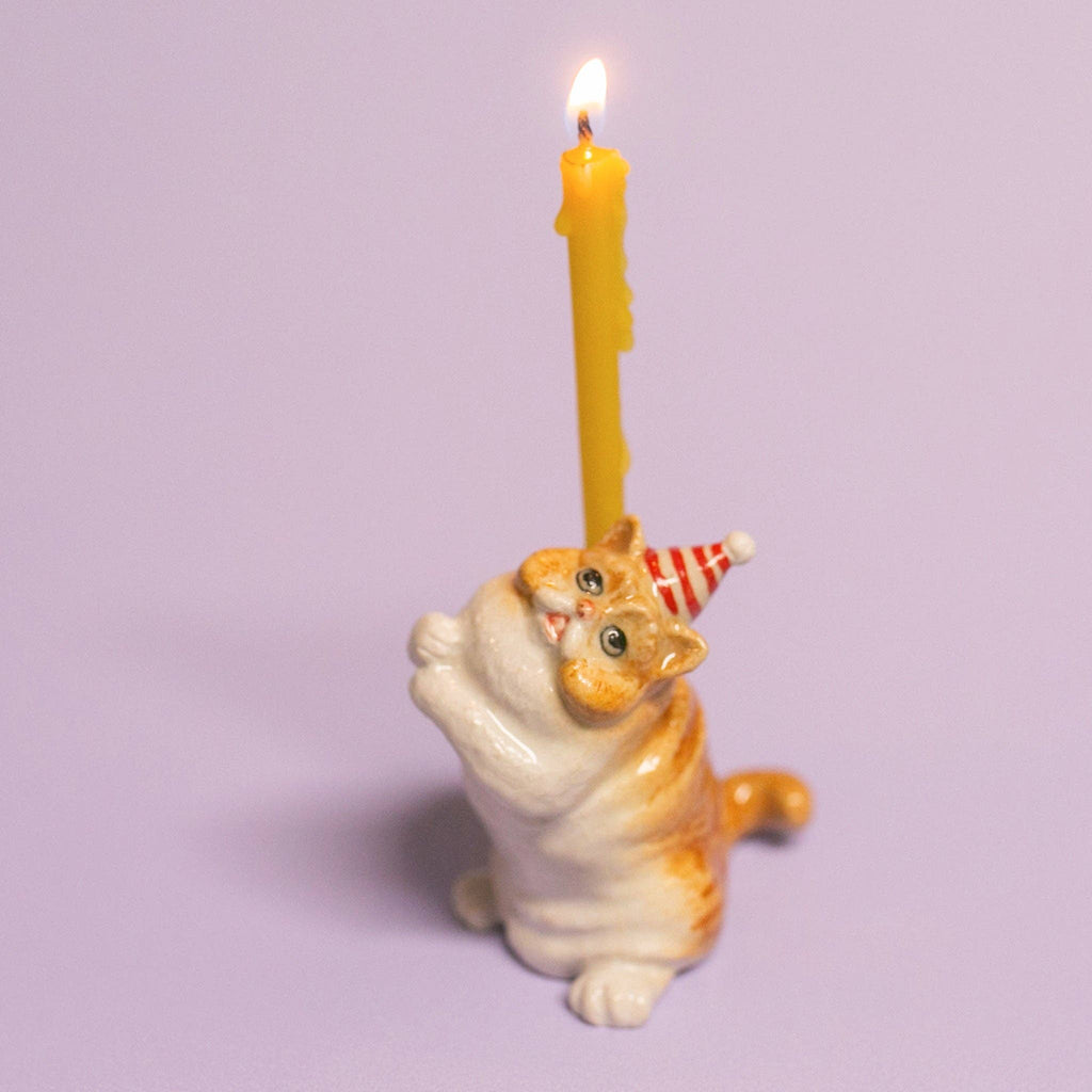 The Exotic Shorthair Cat Cake Topper features a ceramic cat figurine in a red-striped party hat, balancing a lit yellow candle on its back—ideal for cat-themed parties and birthdays. Set against a light purple background.