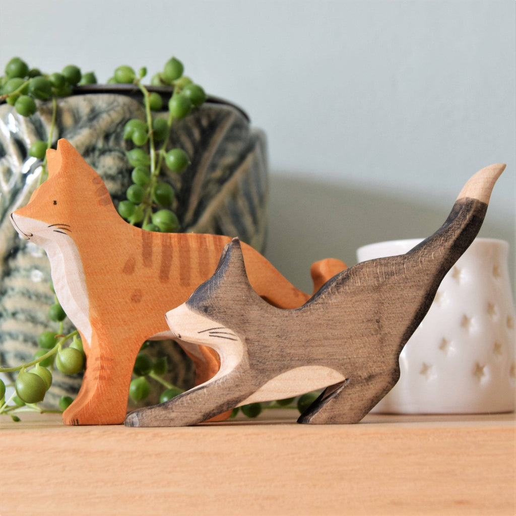 A Handmade Wooden Ginger Cat, crafted from sustainable hardwood, sits on a shelf beside a gray cat figurine. Behind them are a patterned planter with trailing greenery and a white candle holder with star cutouts.