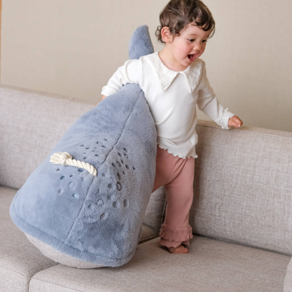 A smiling young child stands on a beige couch, leaning against the Ride-On Whale Pouf—a plush blue pouf featuring a playful whale design and rope handle—radiating happiness and playful energy.