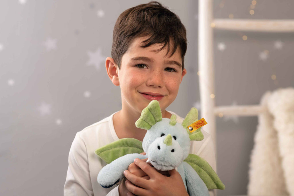 A young boy with short brown hair and freckles smiles while holding a Steiff Samu The Magic Dragon Plush Toy, 11 Inches, in a softly lit room decorated with stars on the gray wall behind him.