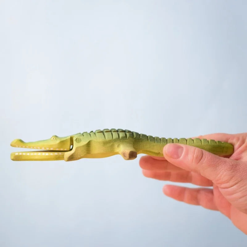 A hand holds a green, crocodile-shaped piece from the Bumbu Wooden Crocodile Cove Set against a light blue background—ideal for Montessori and Waldorf-inspired play.