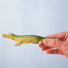 A hand holds a small green crocodile toy from the Bumbu Wooden Crocodile Cove Set, made from sustainably sourced wood, against a plain light blue background.