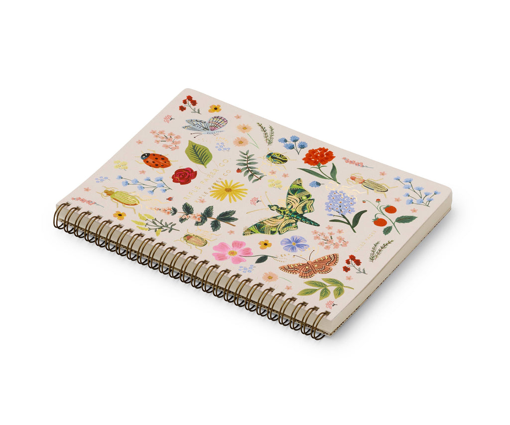 The Curio Spiral Notebook features a spiral binding, a cream cover with colorful flowers, butterflies, and leaves, and ruled pages—ideal for anyone who enjoys charming softcover notebooks.