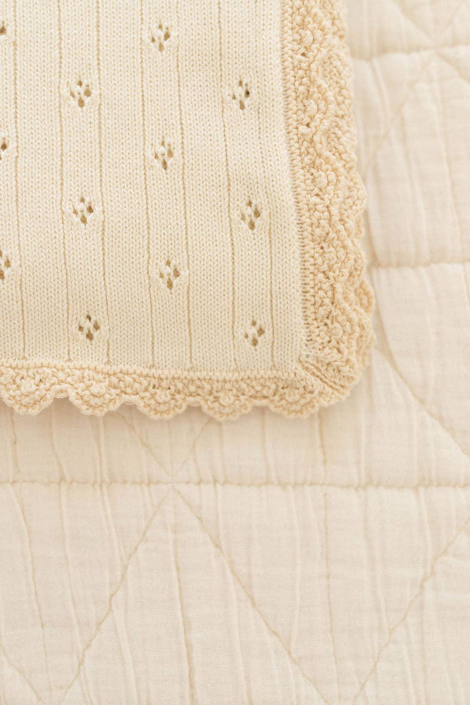 Close-up of the Organic Pointelle Cotton Knit Baby Blanket in cream, crafted in Peru from organic cotton with scalloped edges and heart patterns, displayed on a diamond-quilted fabric surface.