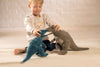 A young child in a light-colored bear shirt sits on the floor, playfully making two Steiff Thaisen Tyrannosaurus "T-Rex" Dinosaur Plush Toys (14") face each other.