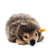 The Steiff Joggi Hedgehog Plush Toy, 6 inches, features brown and beige fur, small black eyes, a black nose, and a yellow Steiff tag on its ear. It is shown lying on a white background.