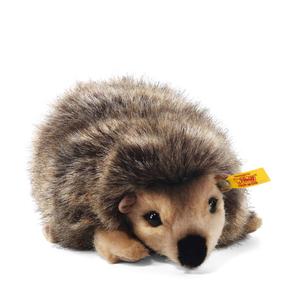The Steiff Joggi Hedgehog Plush Toy, 6 inches, features brown and beige fur, small black eyes, a black nose, and a yellow Steiff tag on its ear. It is shown lying on a white background.