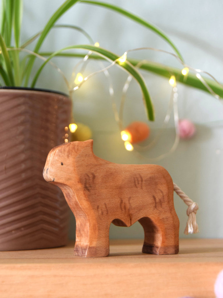The Handmade Wooden Highland Calf, featuring a hand-painted finish and rope tail, stands on a wooden surface. In the background are a patterned plant pot with green leaves and string lights adorned with colorful orbs.