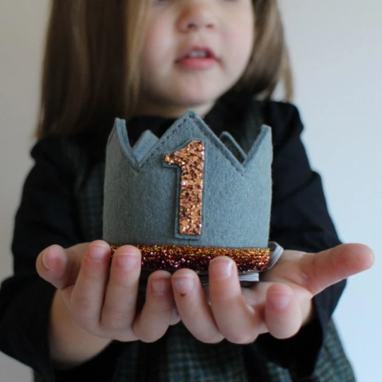 A young child holds out the Storm Gray Felt Crown, ideal for birthdays, adorned with a gold glitter number one and a sparkling band. The child's face is partially visible and softly blurred in the background.