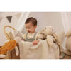 A young child sits in a wicker basket lined with a soft knitted blanket, surrounded by Senger Naturwelt Cuddly Animal - Turtle toys made from organic cotton in a softly lit, neutral-toned room.