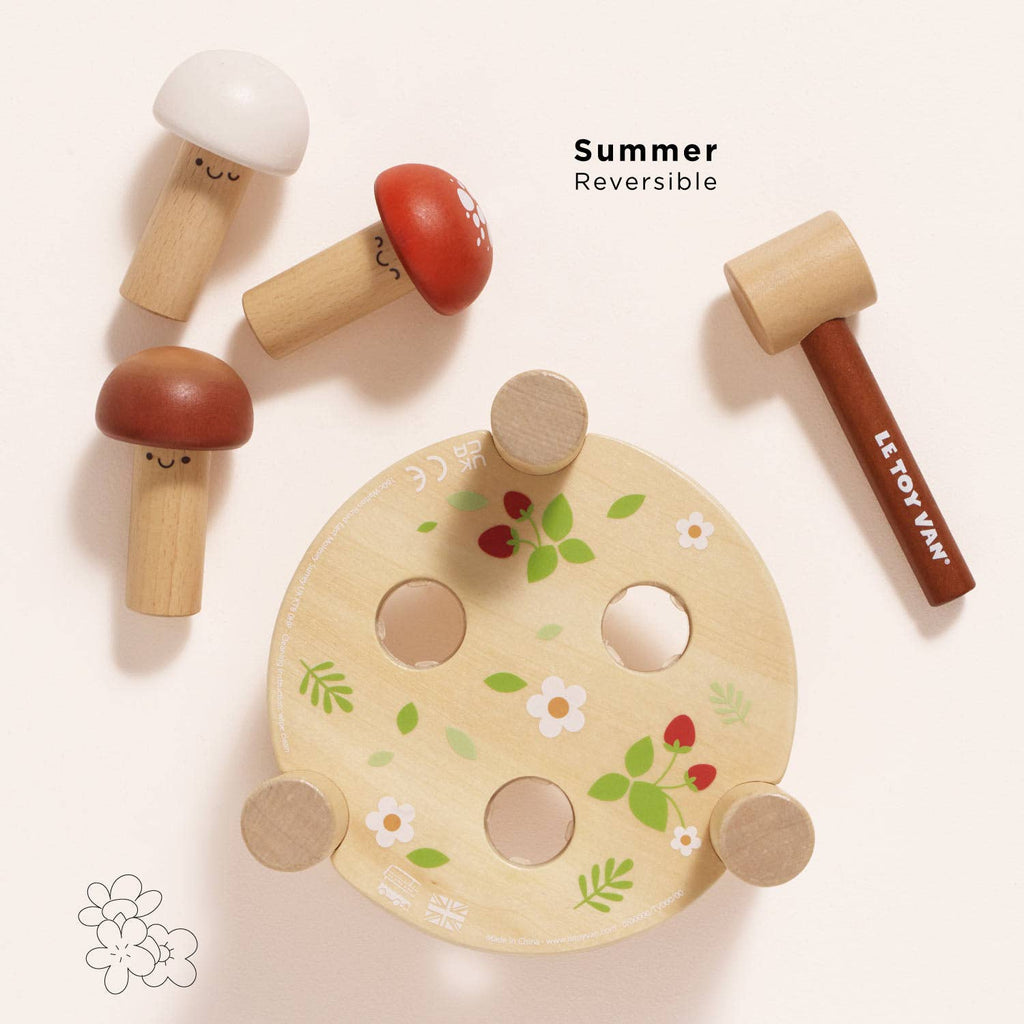 The Woodland Mushroom Hammer Game is a reversible wooden toy with a floral circular base, three smiling mushroom pegs, and a small mallet. Made from FSC-certified wood to encourage fine motor skill development. Text reads "Summer Reversible.