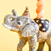 A close-up of the Elephant Cake Topper, a fine porcelain figurine with a striped party hat, star-patterned blanket, and twisted candle on its back, creates an heirloom-quality collectible keepsake against a yellow background.