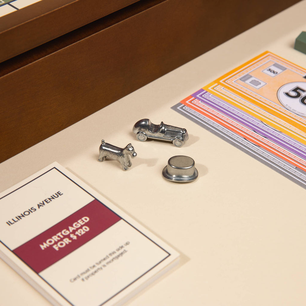 Game pieces—a dog, car, and hat—sit beside property cards, Monopoly money, and a mortgaged Illinois Avenue card, bringing classic charm to the 3-in-1 Game Collection: Monopoly, Sorry & Mancala wooden board game set.