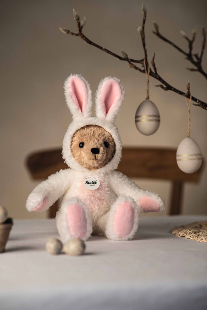 An 11-inch Steiff Teddy Bear with Bunny Hoodie Costume sits on a table, while decorative eggs hang from branches above, creating a festive atmosphere reminiscent of a zodiac lucky charm.