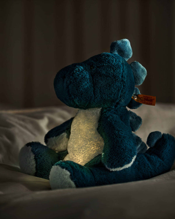 The Steiff Light at Night Spott Stegosaurus Stuffed Plush, 11 inches, sits on a bed softly illuminated by a warm dinosaur night light against a dark, cozy background.