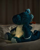 The Steiff Light at Night Spott Stegosaurus Stuffed Plush, 11 inches, sits on a bed softly illuminated by a warm dinosaur night light against a dark, cozy background.