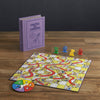 The Chutes and Ladders Vintage Bookshelf Edition features a purple book-shaped box, a vibrant board, spinner, and four colored pieces on a wooden table—perfect for enjoying the classic game’s playful twists and turns.