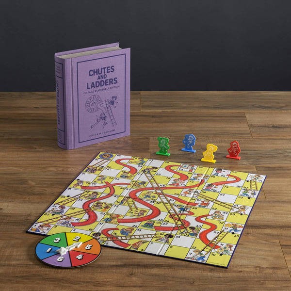 The Chutes and Ladders Vintage Bookshelf Edition features a purple book-shaped box, a vibrant board, spinner, and four colored pieces on a wooden table—perfect for enjoying the classic game’s playful twists and turns.