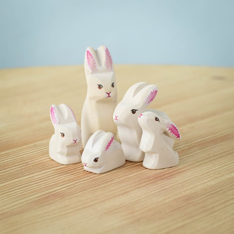 The Bumbu Handmade Wooden White Bunny Rabbit Family Set features five eco-friendly wooden bunnies with pink ears and black eyes, arranged on a light wood surface against a soft blue background—ideal for Montessori toy collections.