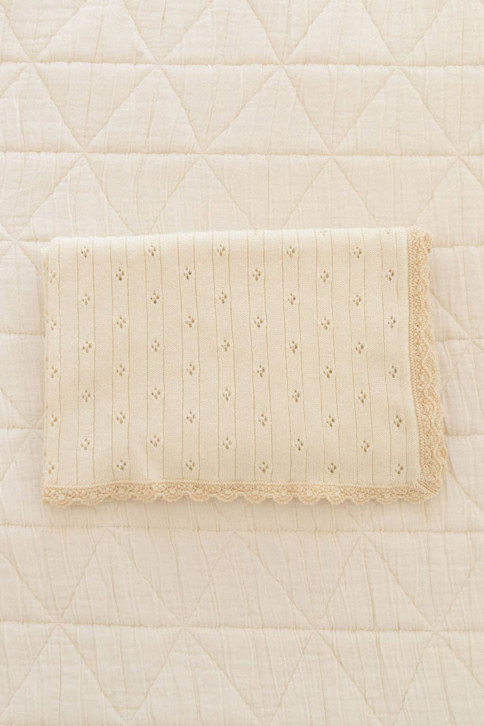 The Organic Pointelle Cotton Knit Baby Blanket, cream-colored with a scalloped lace edge and decorative holes, is beautifully made in Peru and shown folded on a quilted diamond-pattern cream bedspread.