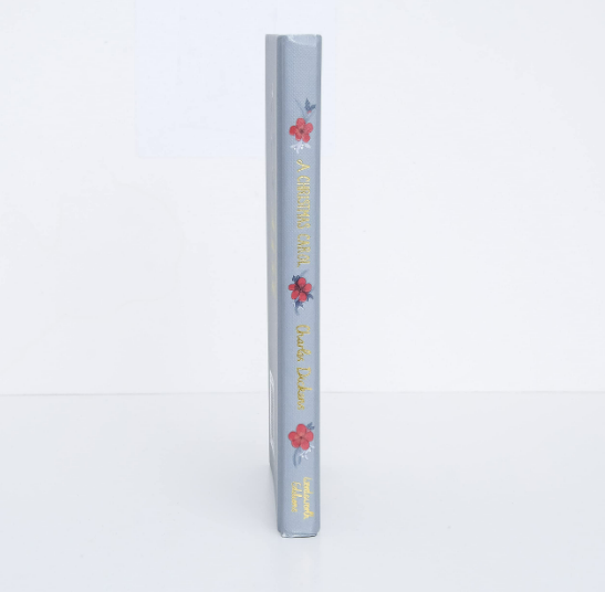 A light gray hardcover book standing upright with floral designs on the spine, featuring red flowers and green leaves. The spine reads "A Christmas Carol | Collector's Edition| Hardcover Book.