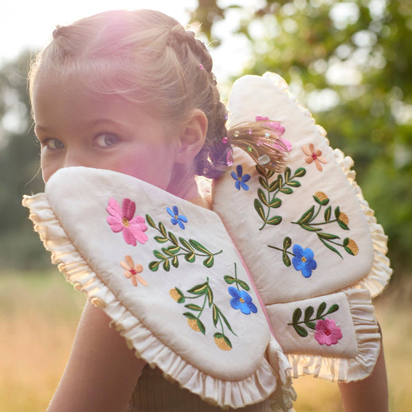 A young girl with braided hair stands outdoors in sunlight, wearing Meri Meri Butterfly Wings crafted from soft, natural cotton and embroidered with colorful flowers, surrounded by greenery.