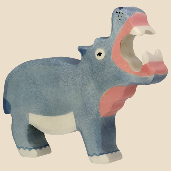 The Holztiger Hippo, a handcrafted and hand-painted blue maple and beech wood figurine, stands on all fours with an open mouth showing pink and white details. Inspired by classic HOLZTIGER figures. Ships in one week. Set against a beige background.