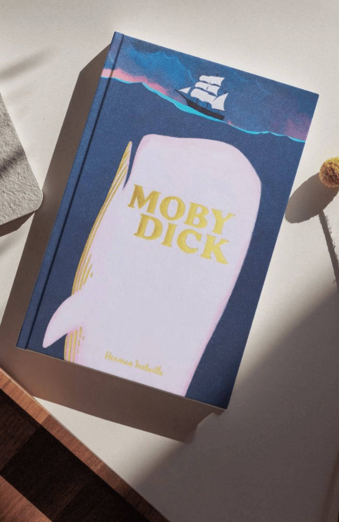 The "Moby Dick | Melville | Collector's Edition | Hardcover," with a pink whale and sailing ship on the cover, rests on a sunlit white surface.