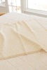 The Organic Pointelle Cotton Knit Baby Blanket, in a soft cream hue with a subtle dotted pattern and scalloped edges, is draped neatly over a quilted bed in a bright, sunlit room.